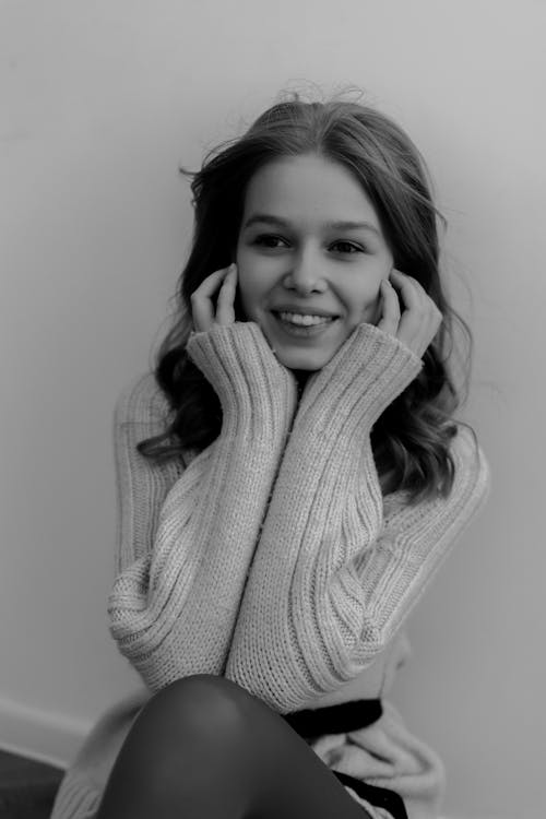Black and White Photo of a Young Woman in a Sweater Sitting by the Wall and Smiling 