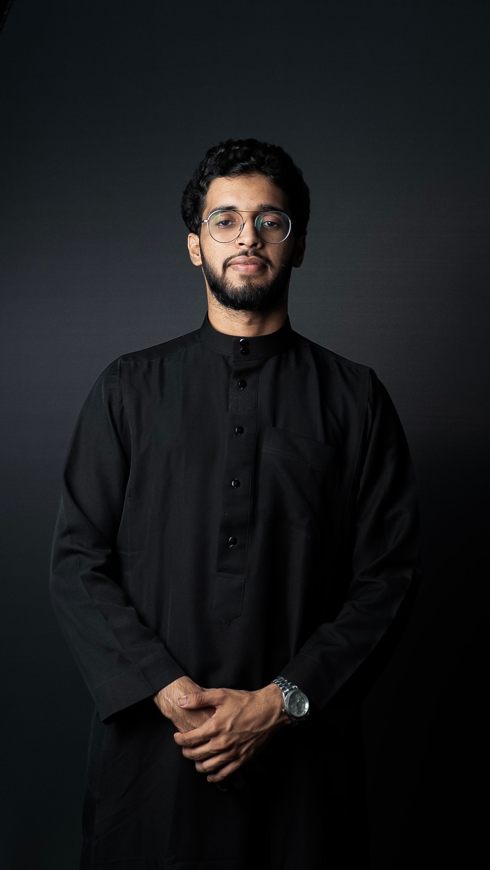 Elegant portrait of a man wearing traditional black clothing with glasses, set against a dark background.