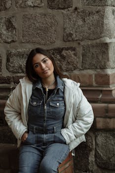Stylish woman in denim and jacket leaning against a rustic stone wall.