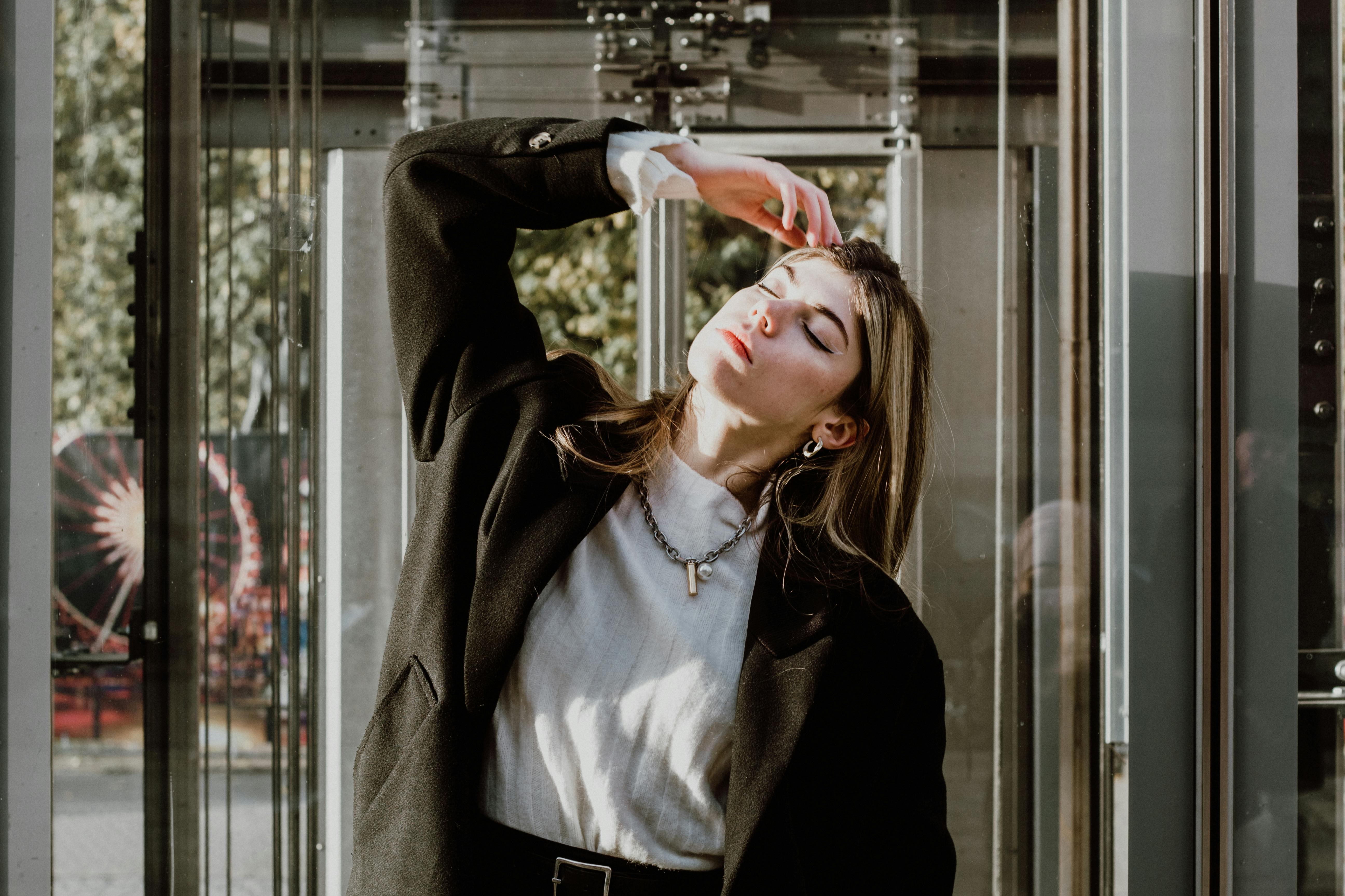 A fashionable woman poses confidently in sunlight by a glass building in the city.