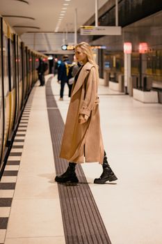 A blonde woman in a coat walking through a modern urban subway station.