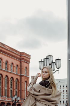 A fashionable woman with blonde hair in an elegant coat walking through a city street lined with historic buildings during the fall season.