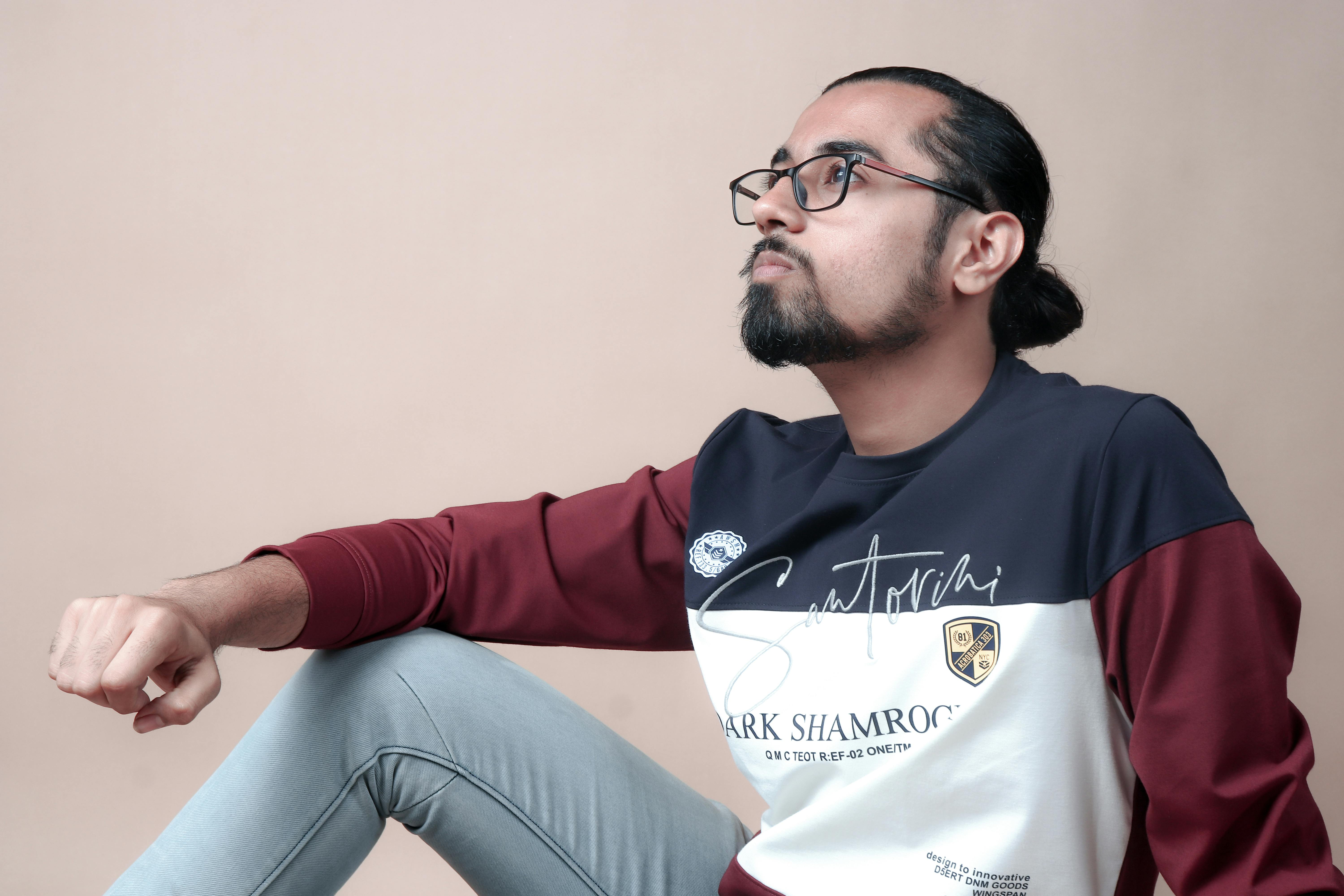 A fashionable bearded man with eyeglasses sits thoughtfully, wearing a trendy outfit.