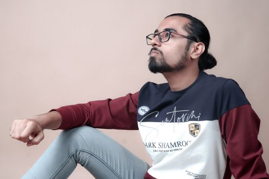 A fashionable bearded man with eyeglasses sits thoughtfully, wearing a trendy outfit.