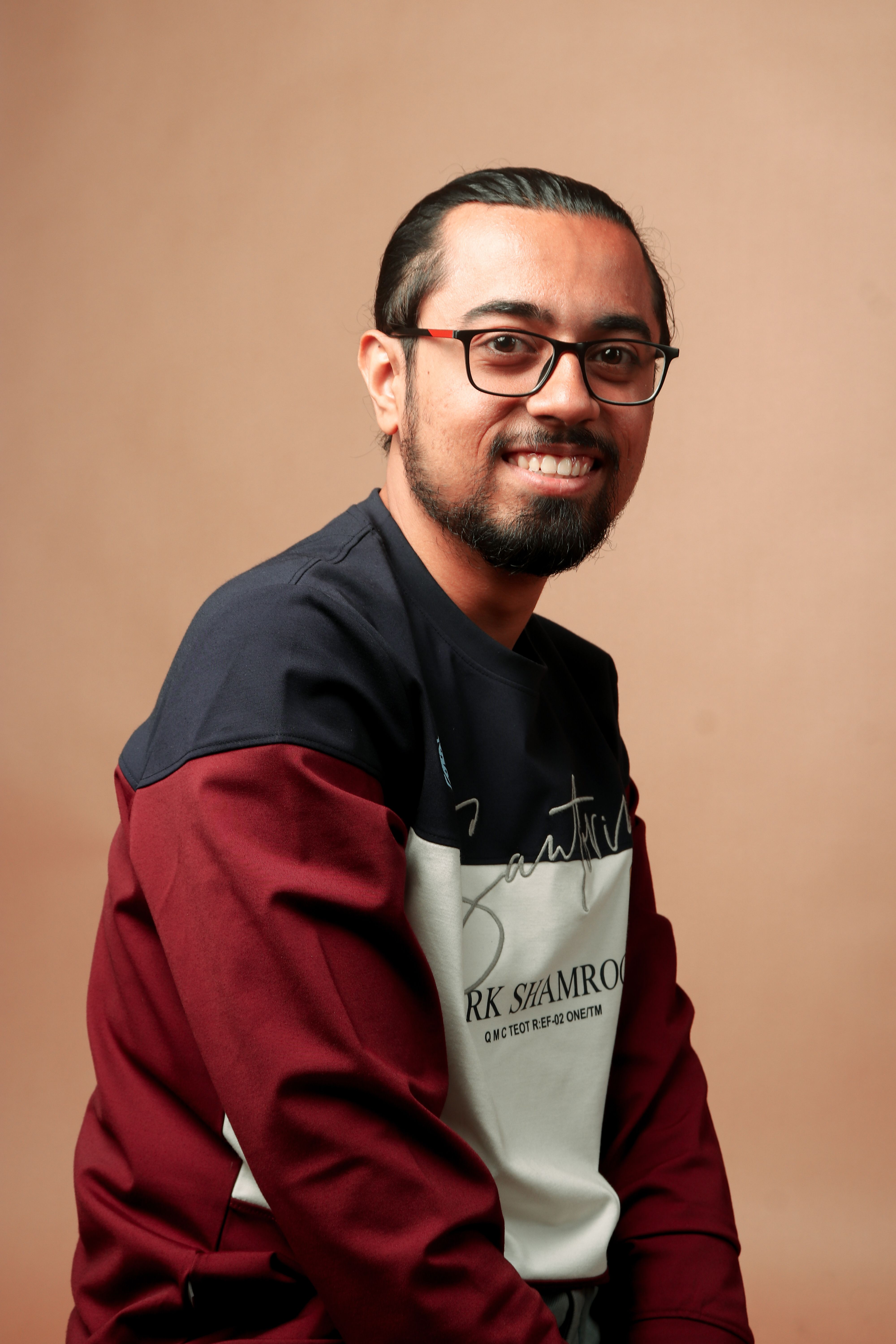 Portrait of a smiling man with eyeglasses and a beard in a casual outfit, studio setting.