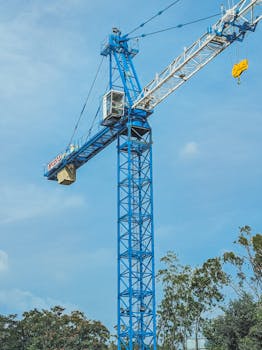 A towering blue construction crane stands tall against a clear blue sky, surrounded by trees.