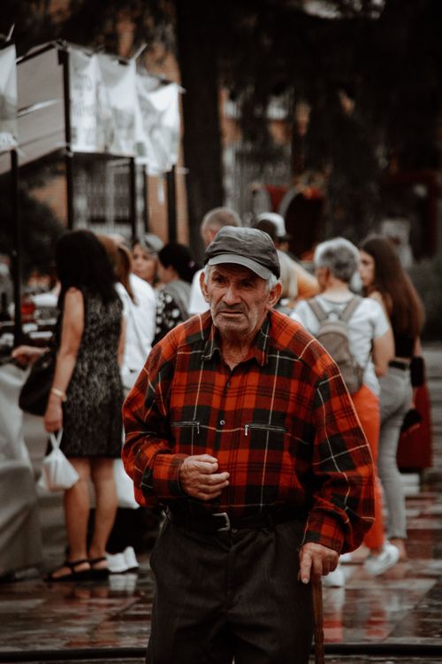Free An elderly man in a plaid shirt walks through a bustling outdoor market. Stock Photo