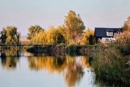Free A peaceful rural scene with a lake, footbridge, and rustic cabin surrounded by autumn trees. Stock Photo