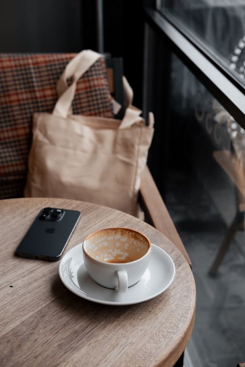 Free A warm coffee setting with a latte, smartphone, and bag by a window in a cozy cafe. Stock Photo