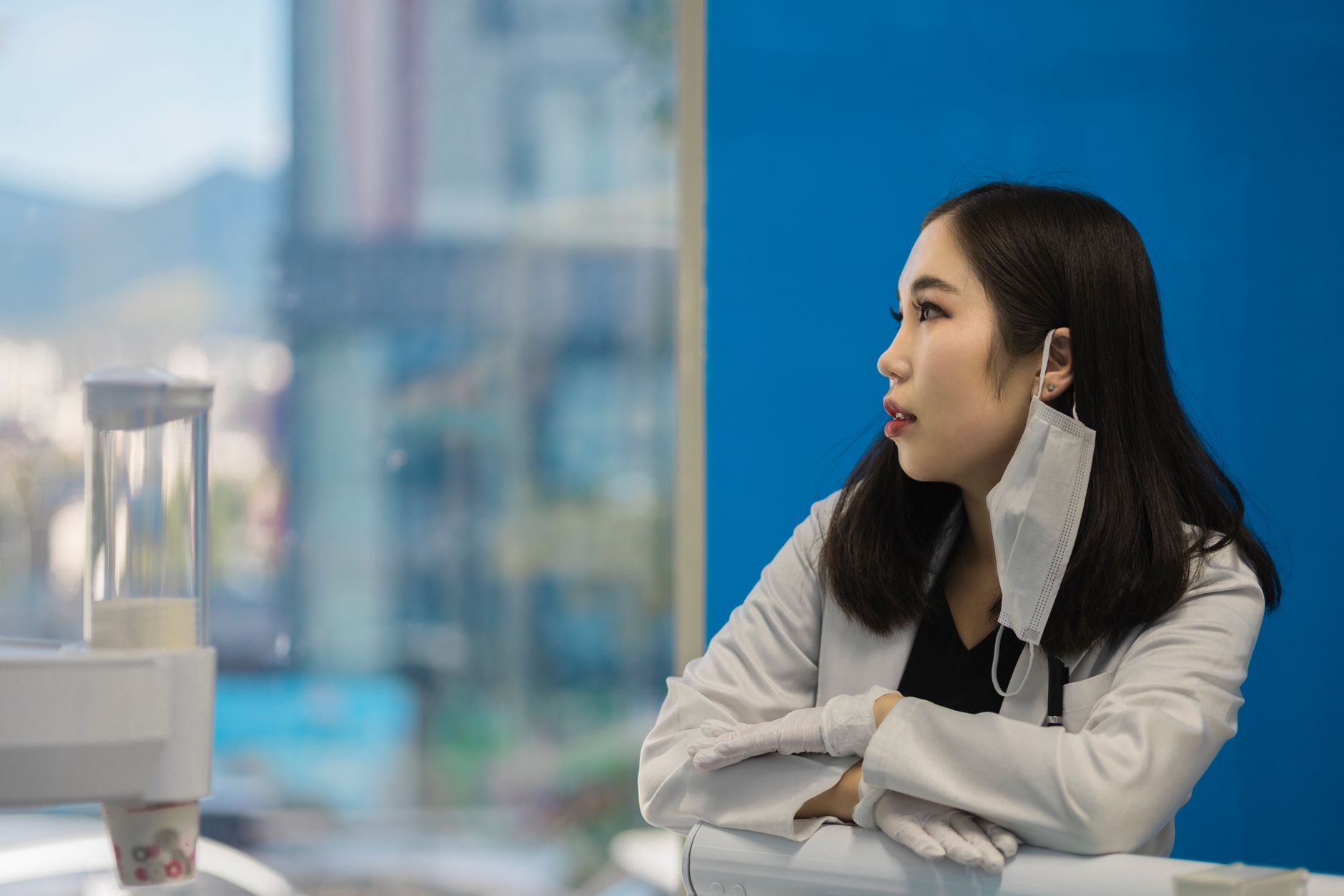 Asian female scientist in lab coat with mask and gloves, gazing outside a window.