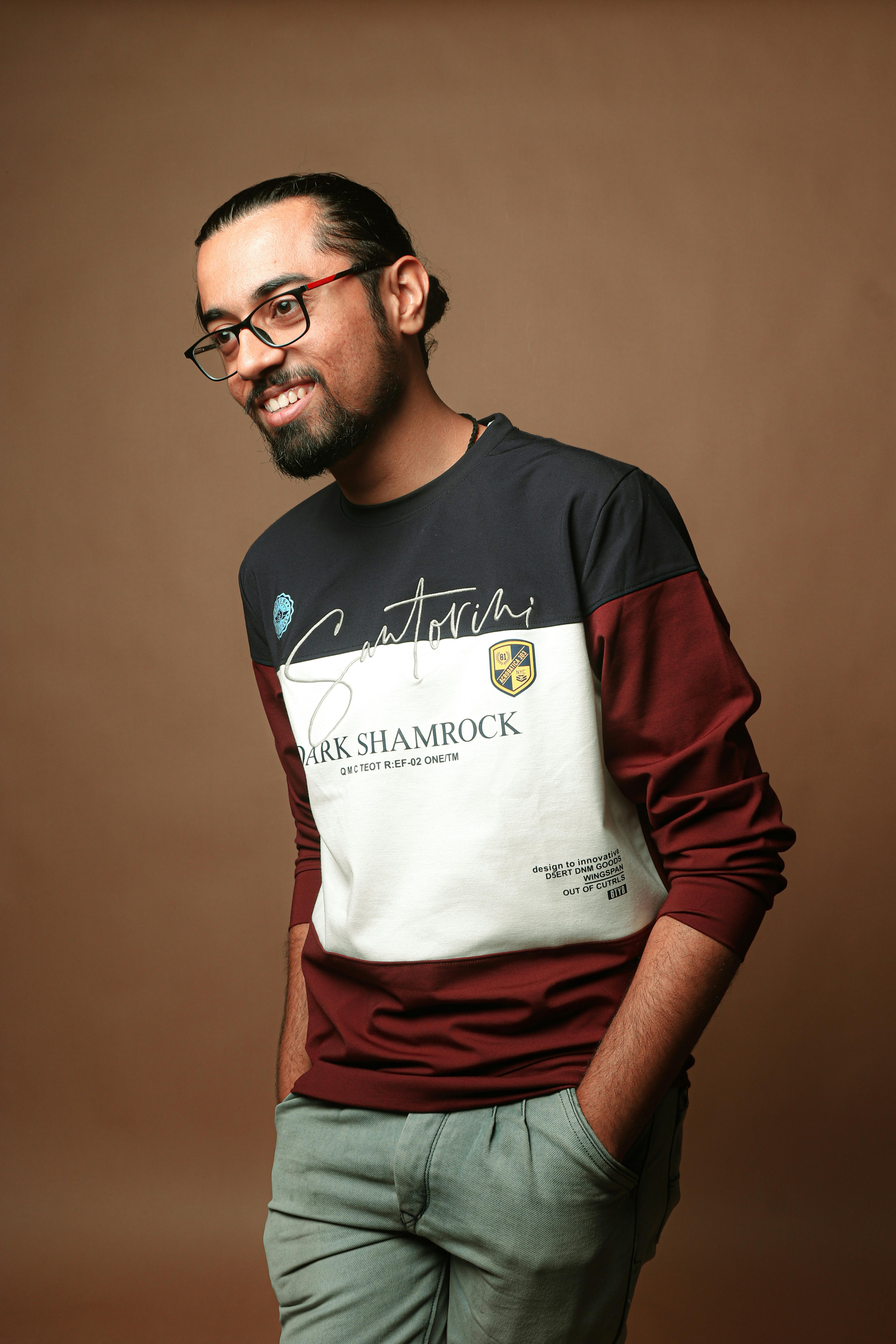 Happy man with beard and glasses posing in studio, wearing casual outfit with hands in pockets.