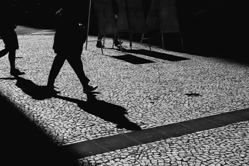 Free Dynamic black and white street photo capturing silhouettes and shadows of people walking on cobblestone pavement. Stock Photo