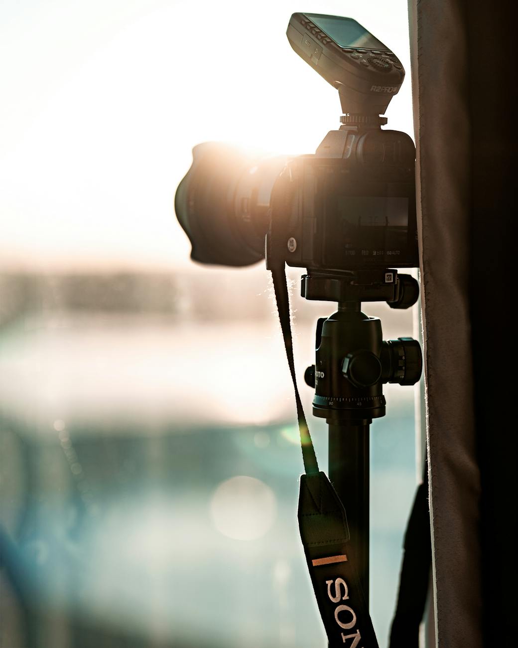 A DSLR camera with a flash setup on a tripod, backlit by warm sunlight, suggesting a professional photography scene.