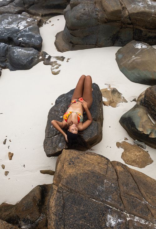 Free Woman in colorful bikini lounging on rocks by the sandy shore, captured in a high angle view. Stock Photo