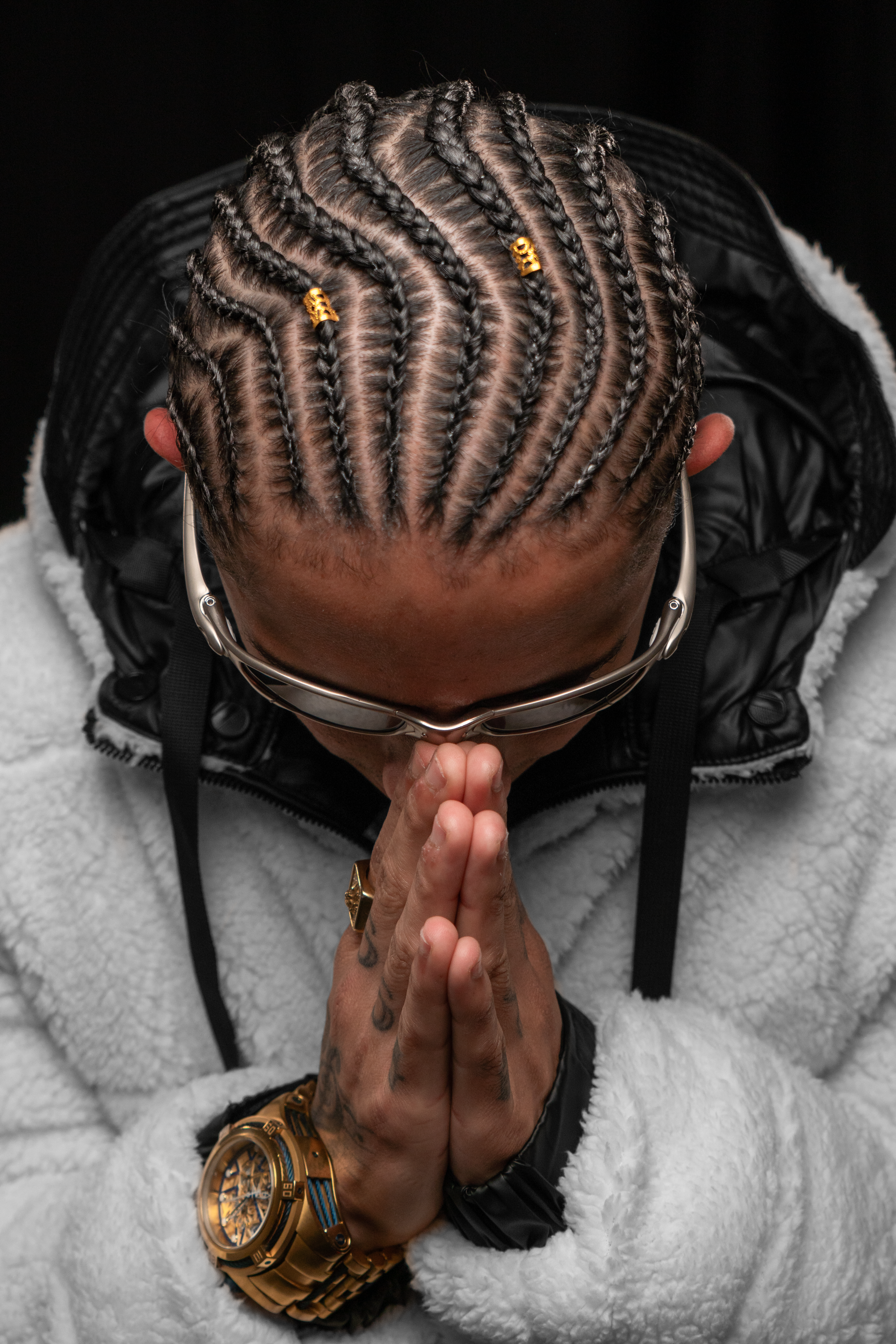 Man with braided hair and sunglasses clasping hands in a reflective pose.