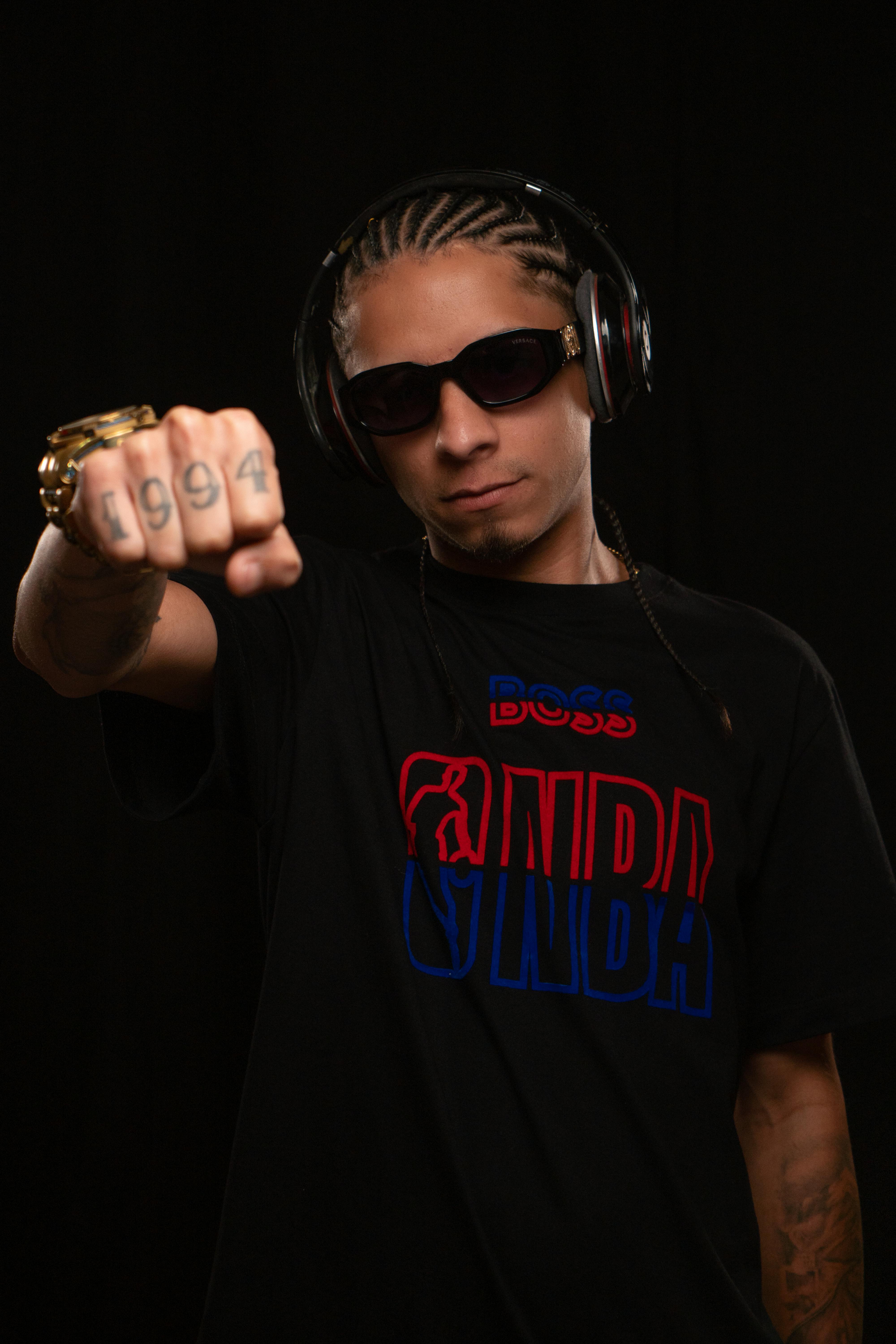 Man in sunglasses with headphones and tattooed fist posed against a black background.
