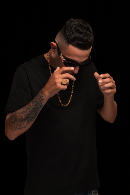 Fashion portrait of a man with sunglasses, gold chain, and black outfit on a dark backdrop.