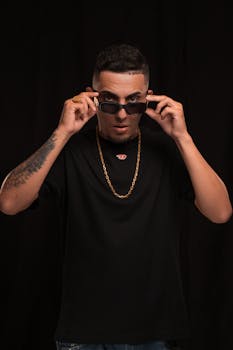 Stylish man in black t-shirt adjusting sunglasses against a dark background.