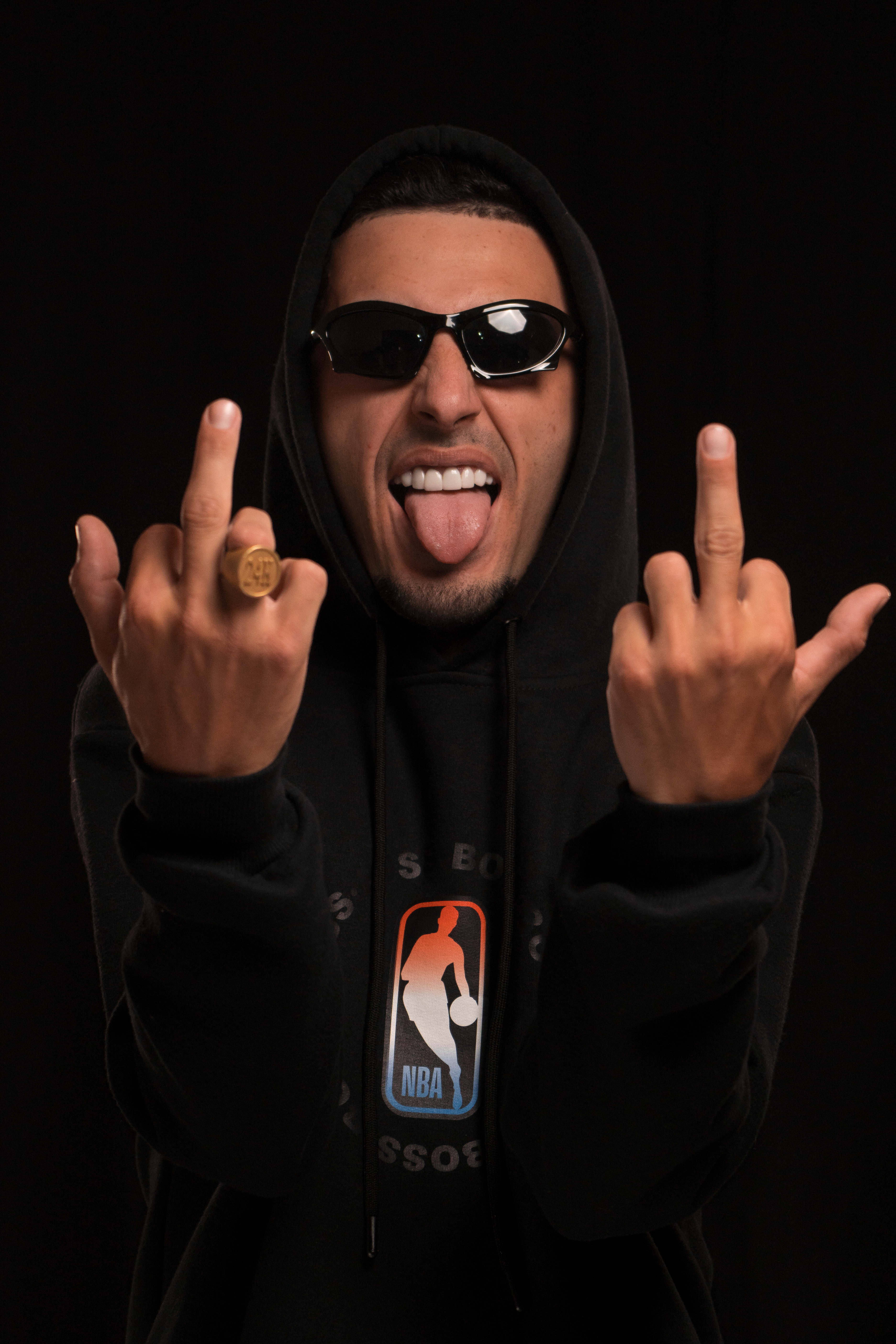 Man in hoodie gesturing boldly in studio setting with sunglasses and ring.