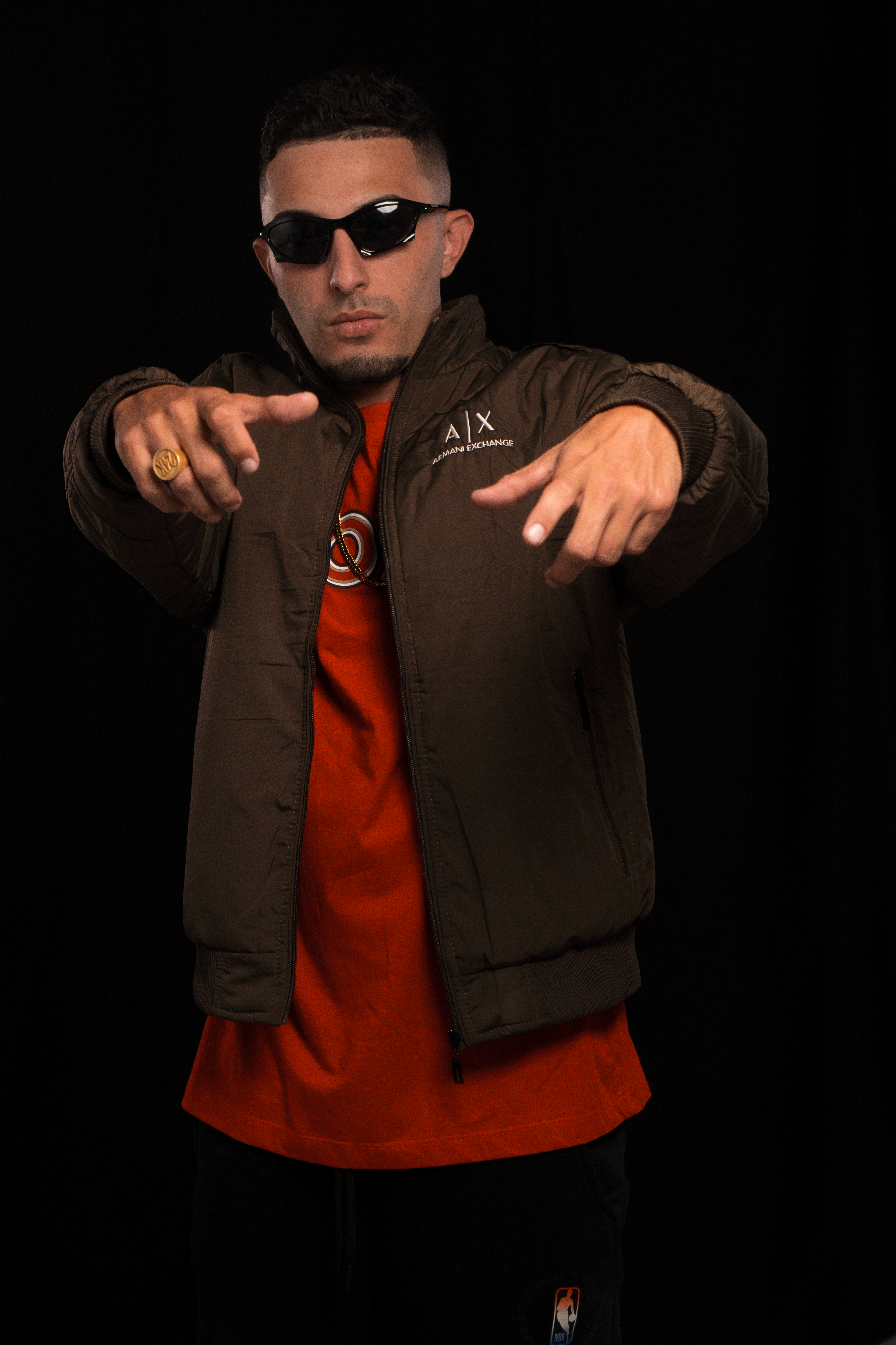Portrait of a man wearing sunglasses and a stylish jacket posing confidently against a black background.