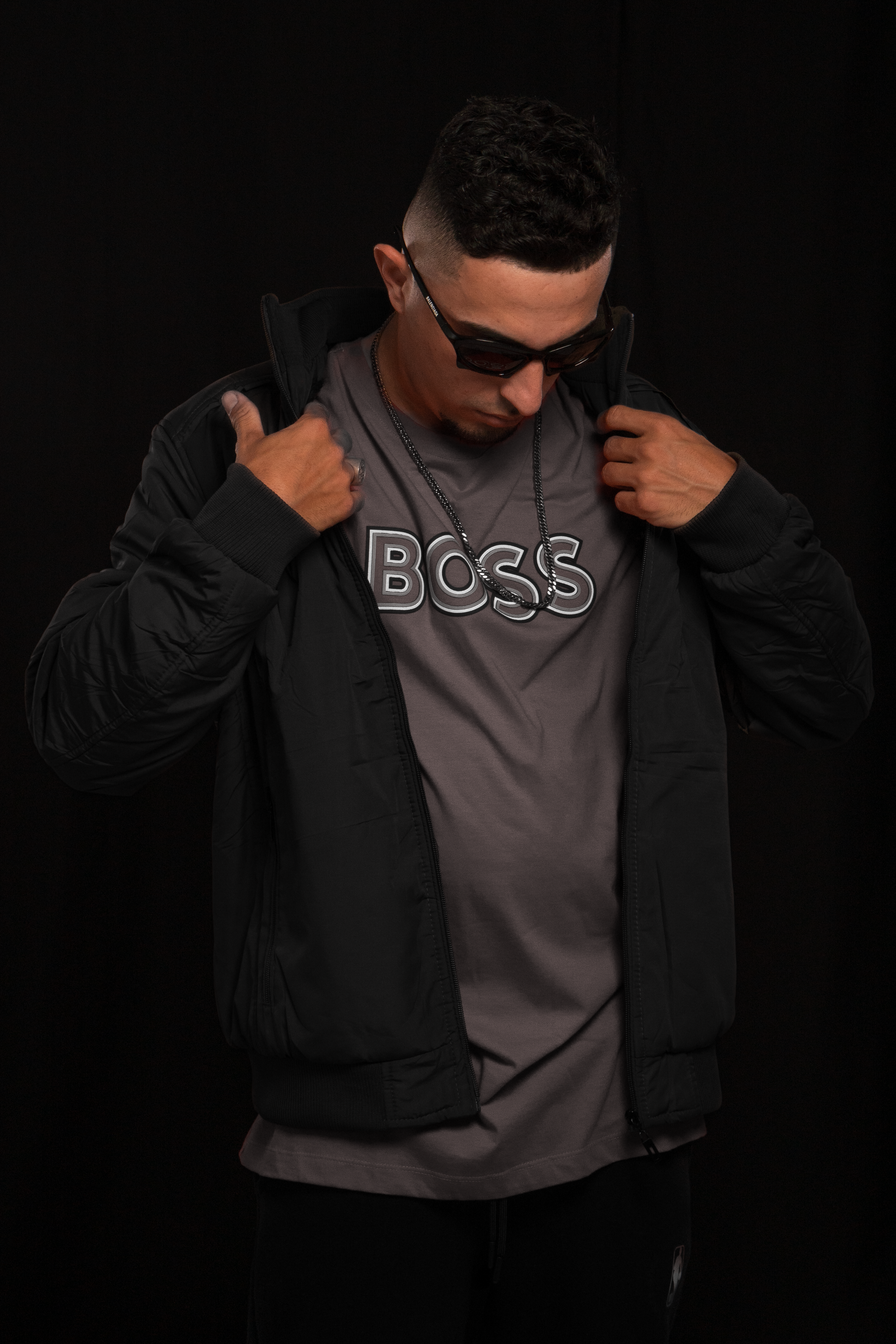 Portrait of a man in sunglasses and jacket against a black backdrop, exuding confidence and style.
