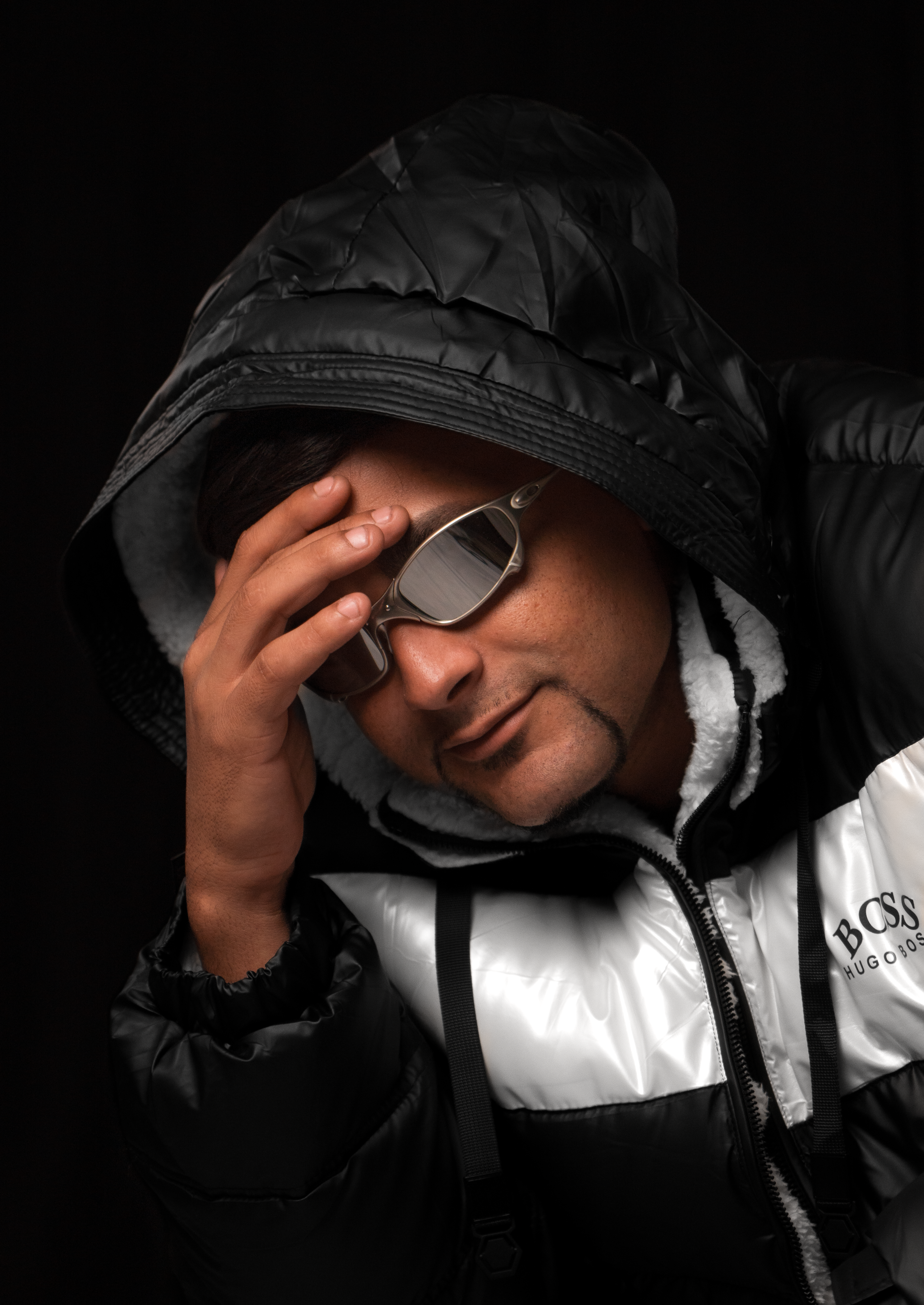 Fashionable man wearing a hooded jacket and sunglasses striking a modern pose against a dark background.