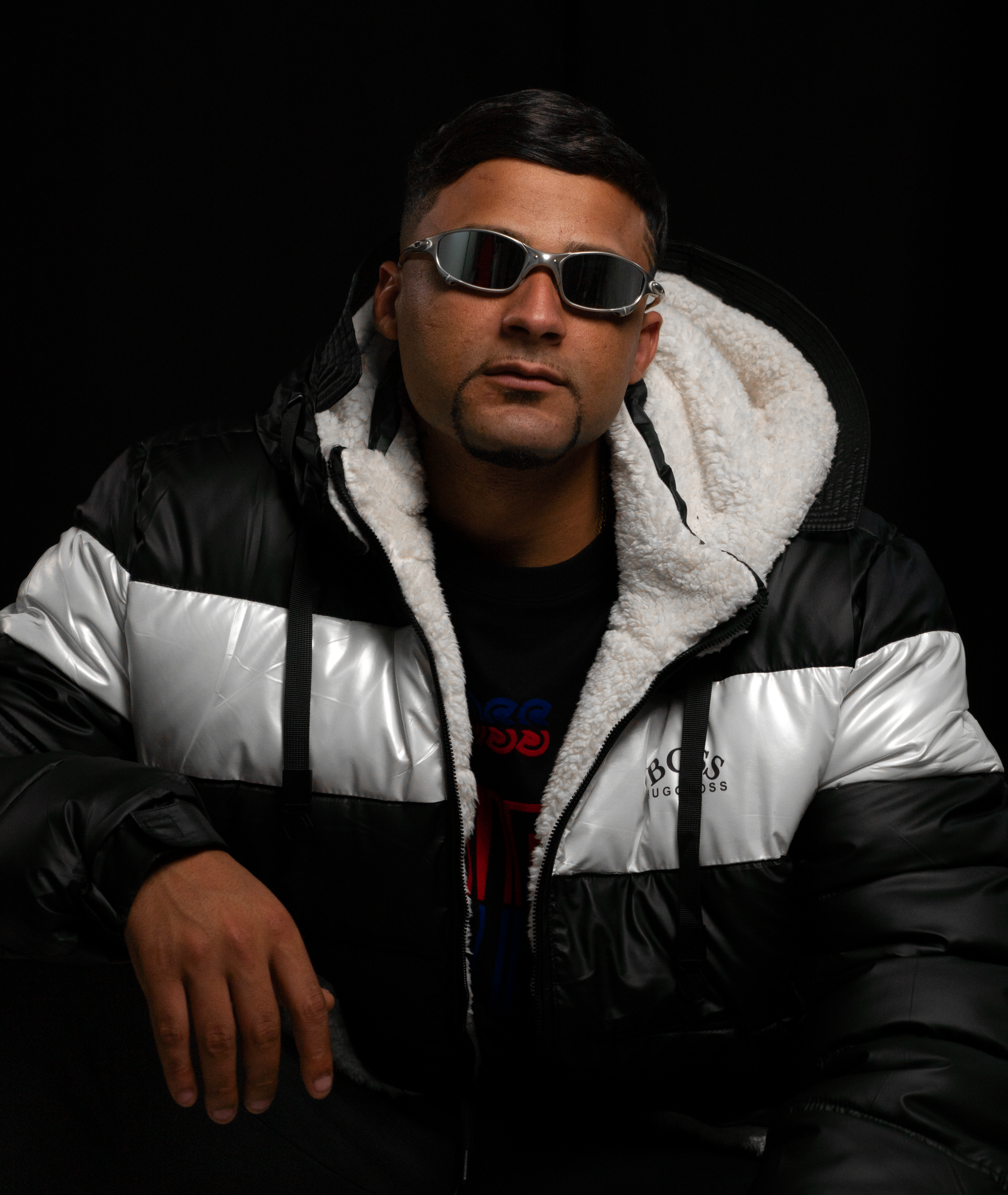 Portrait of a man in a black and silver jacket, wearing sunglasses against a dark background.