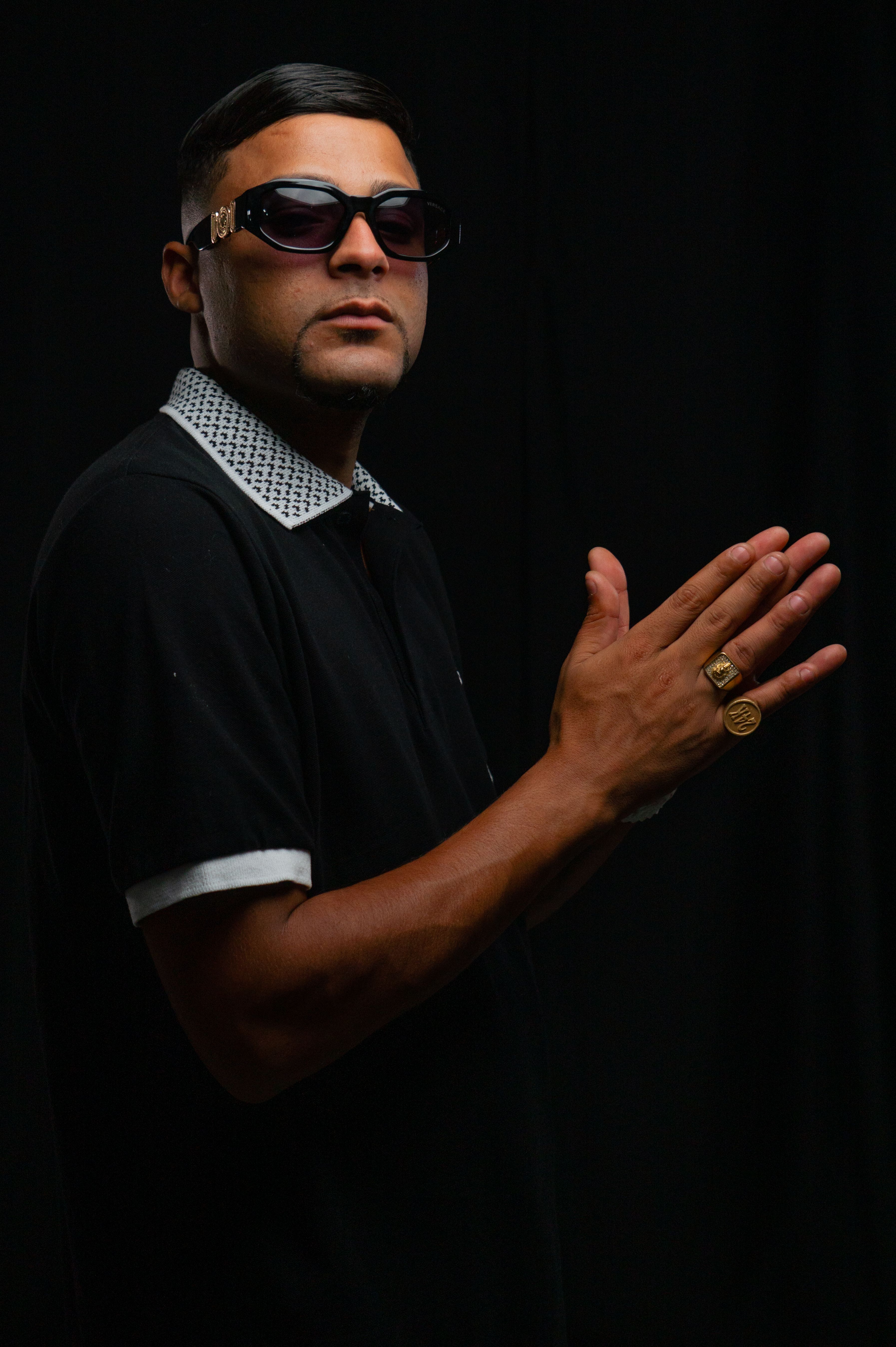 Portrait of a man with sunglasses and clasped hands against a dark backdrop.