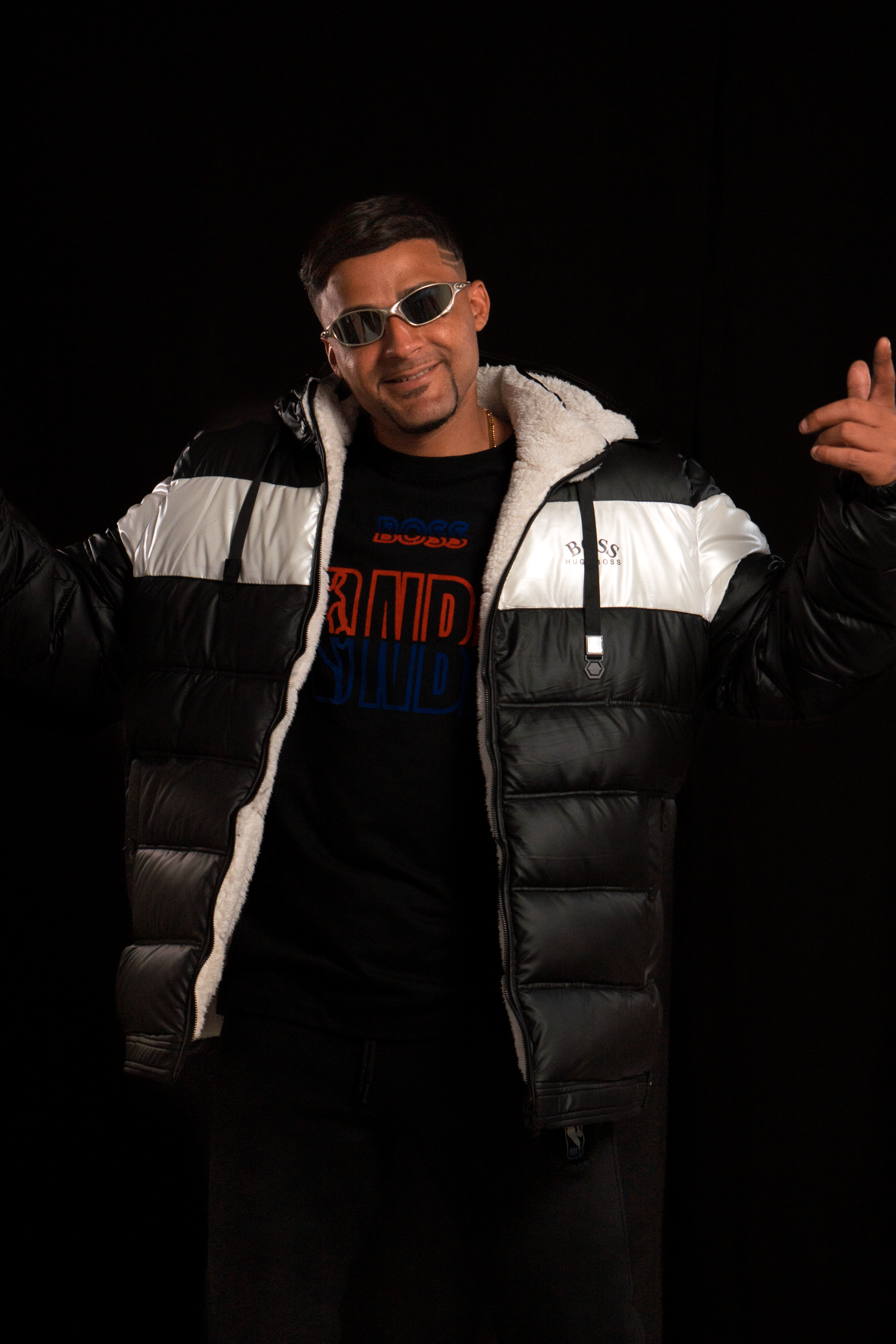 Stylish man in sunglasses and jacket poses confidently against black background.