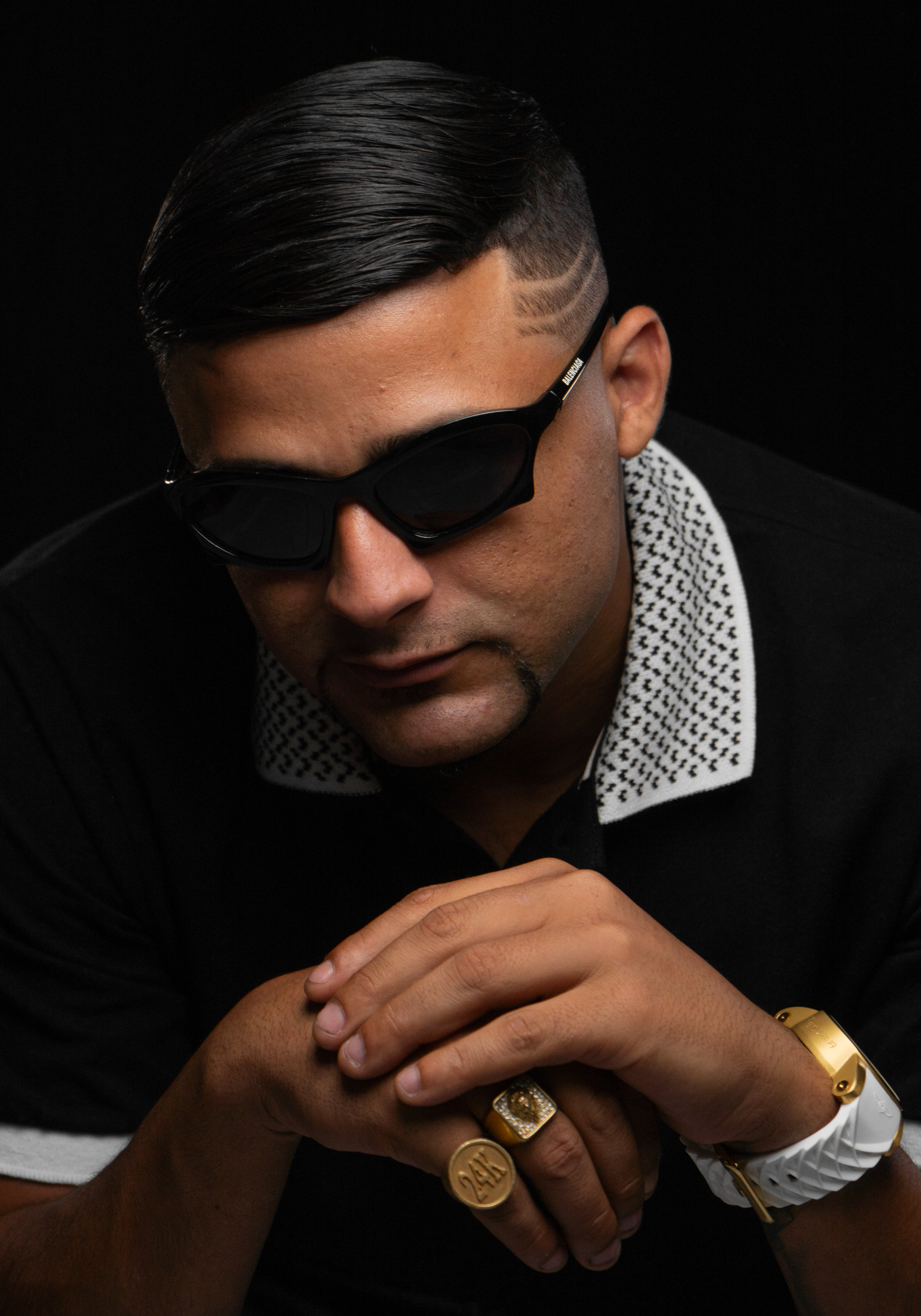 Portrait of a man with sleek hairstyle and jewelry, wearing sunglasses on a dark backdrop.