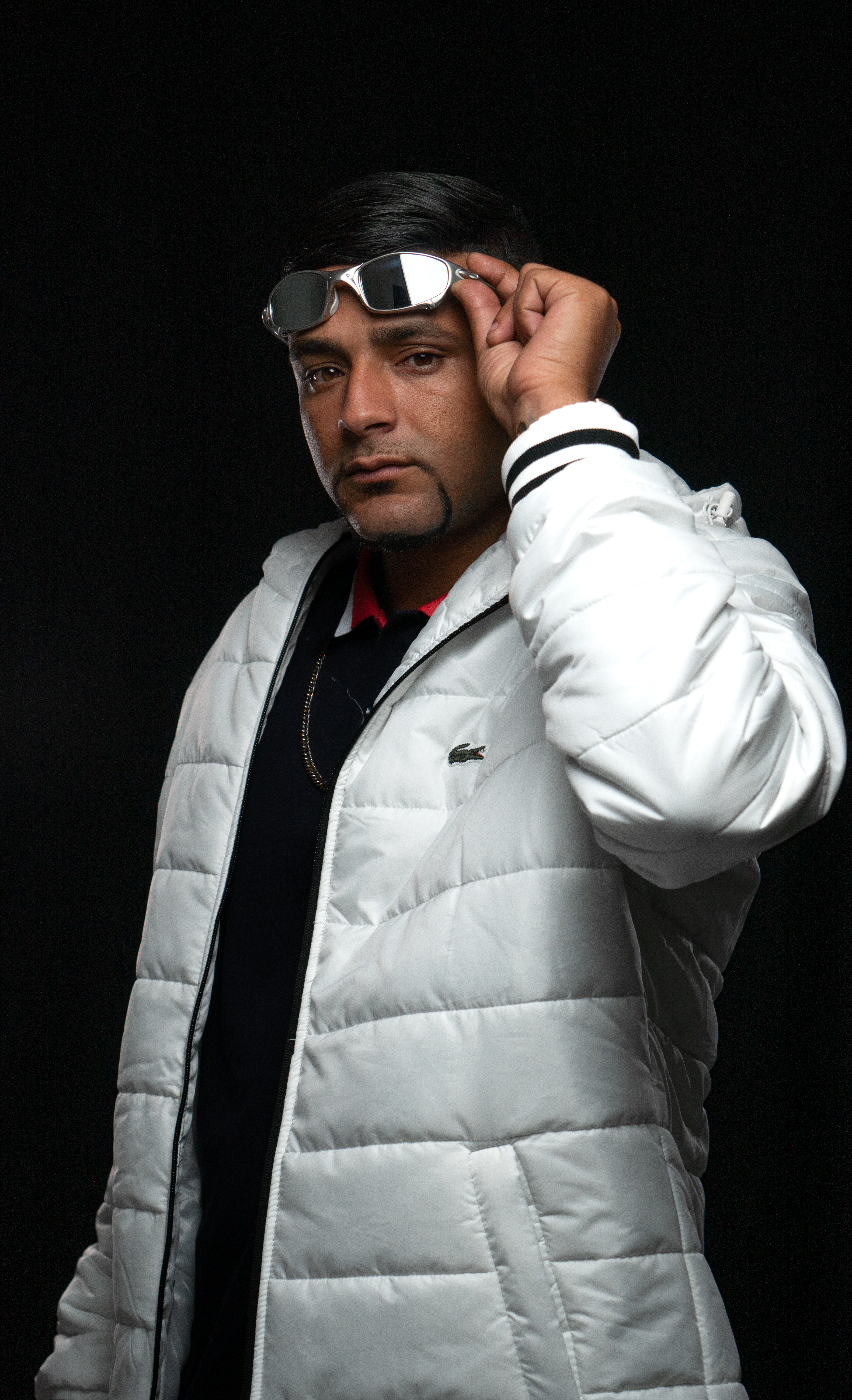 Fashion portrait of a man in a white jacket adjusting sunglasses against a black background.