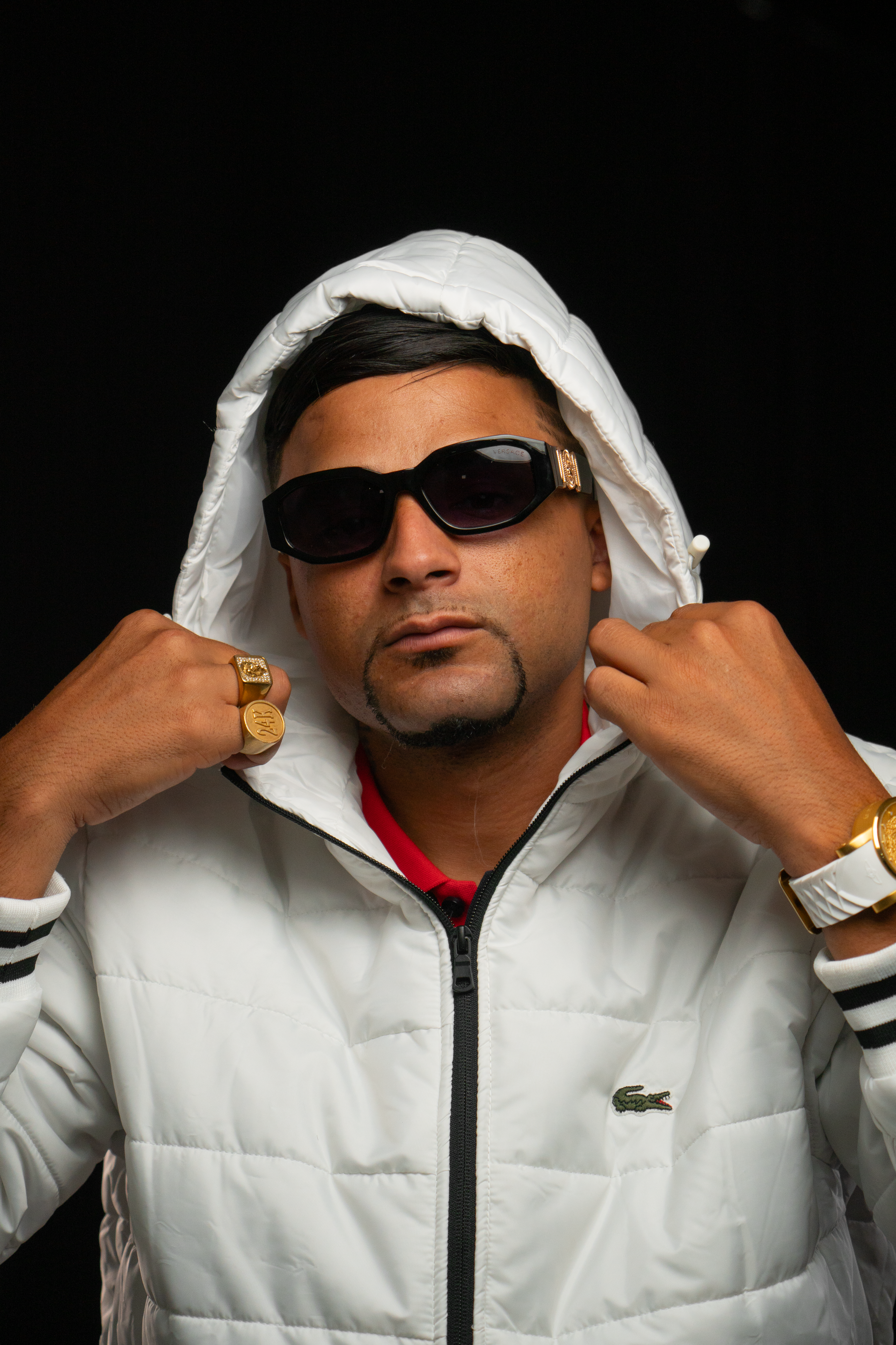 Portrait of a fashionable man in sunglasses and jewelry wearing a white hooded jacket against a black background.