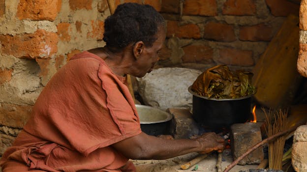 An elderly African woman cooks traditional food over an open fire in a rustic kitchen.