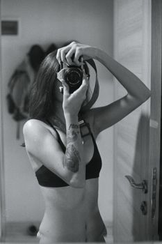 Artistic black and white portrait of a tattooed woman capturing a mirror selfie with a camera.