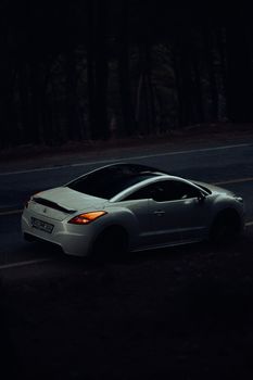 Modern white car driving on a winding road through a forest at twilight in Fethiye, Türkiye.