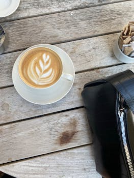Cup of cappuccino with latte art on a wooden table beside a black handbag.