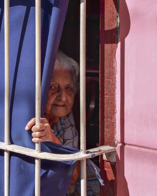 Free An elderly woman gazes through barred window curtains, capturing curiosity and warmth. Stock Photo