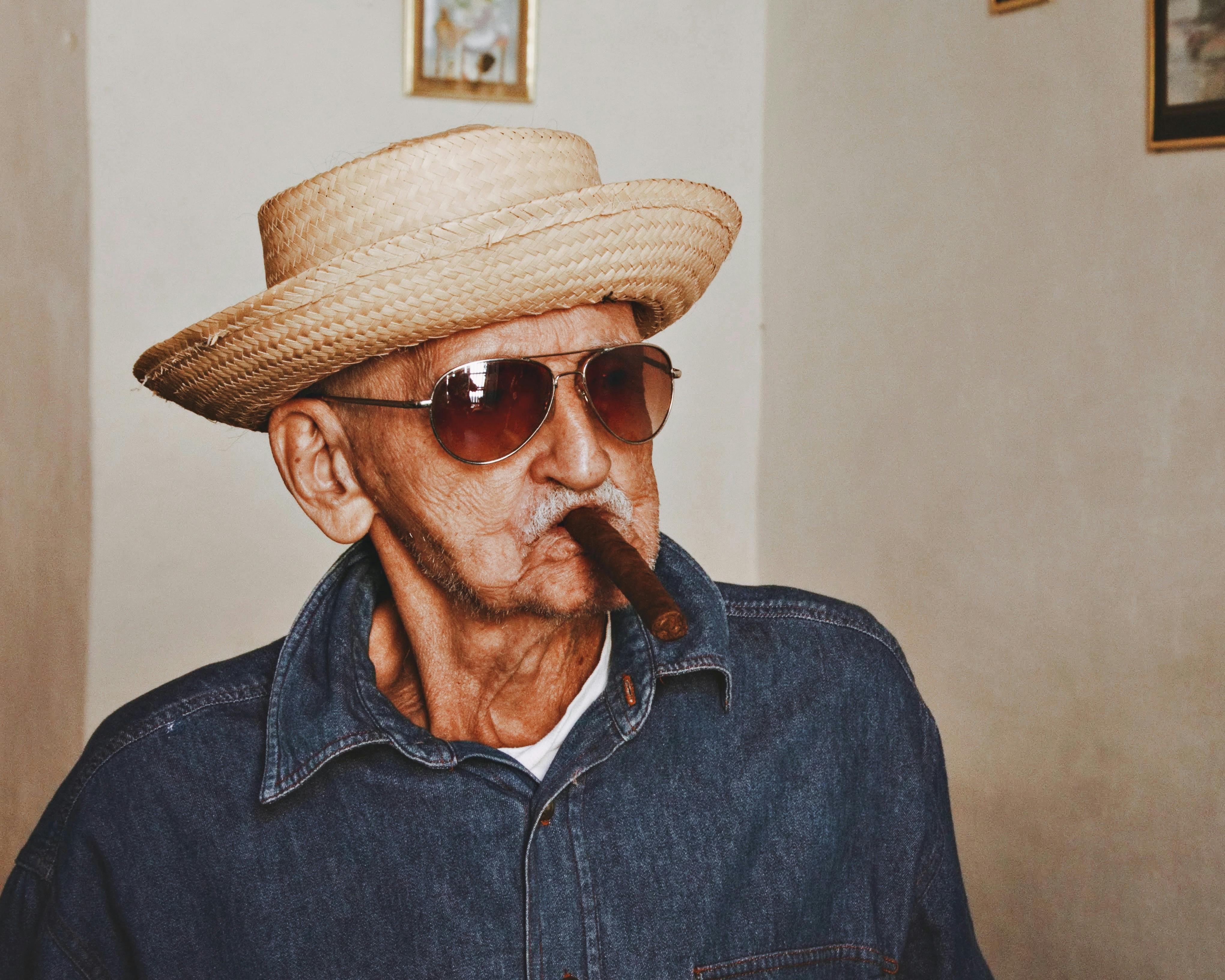 Senior man with sunglasses and straw hat smoking a cigar indoors.