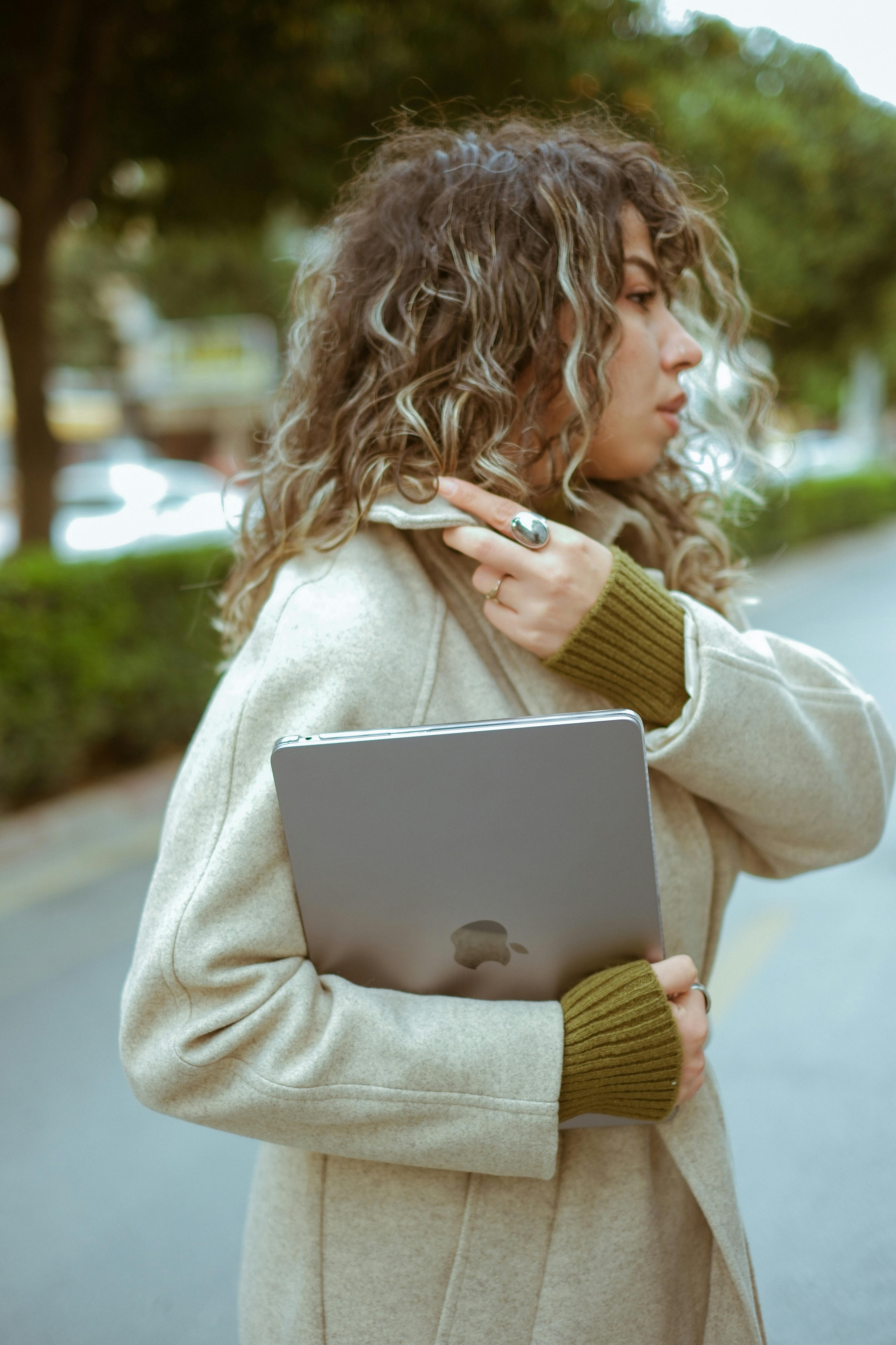 Stylish woman with curly hair walking on a city street holding a laptop.
