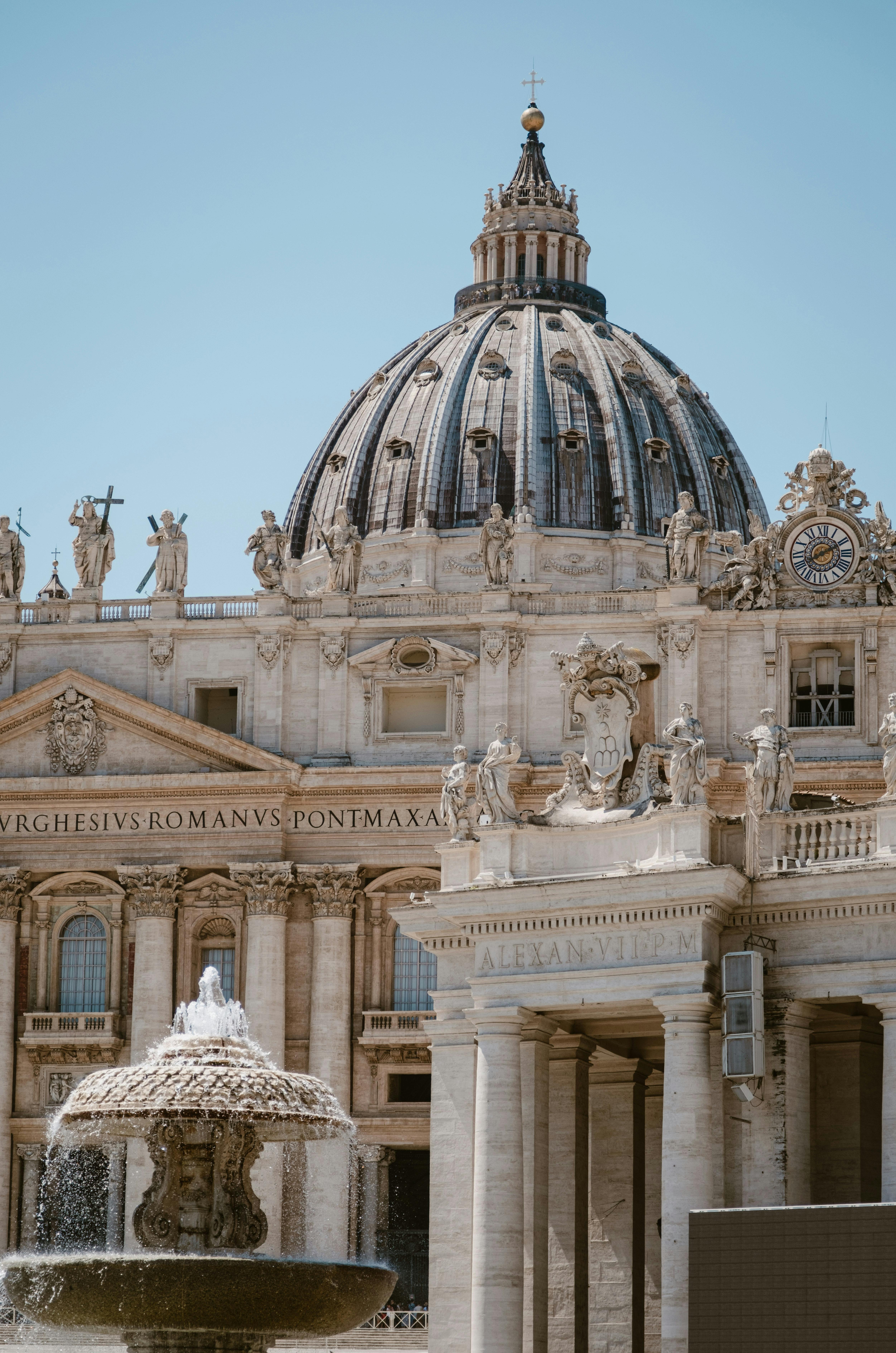 Capture of St. Peter's Basilica dome and a fountain under clear blue skies in Vatican City.