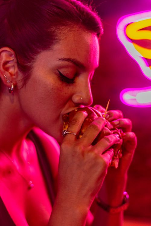 Free A woman savoring a burger under dynamic neon lights, highlighting a vibrant nightlife atmosphere. Stock Photo