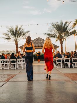 Two women in elegant gowns walking at an outdoor event under palm trees.