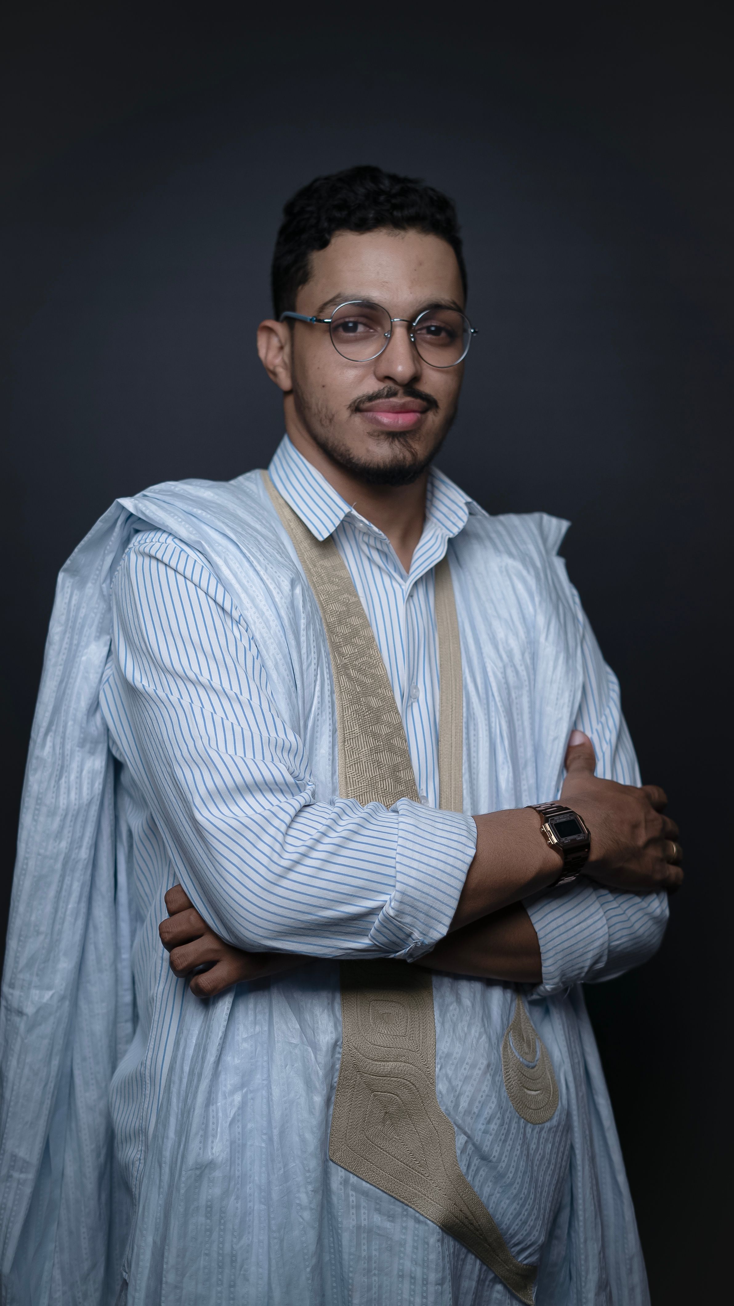 Portrait of a young man in traditional Mauritanian attire with arms crossed against a gray background.
