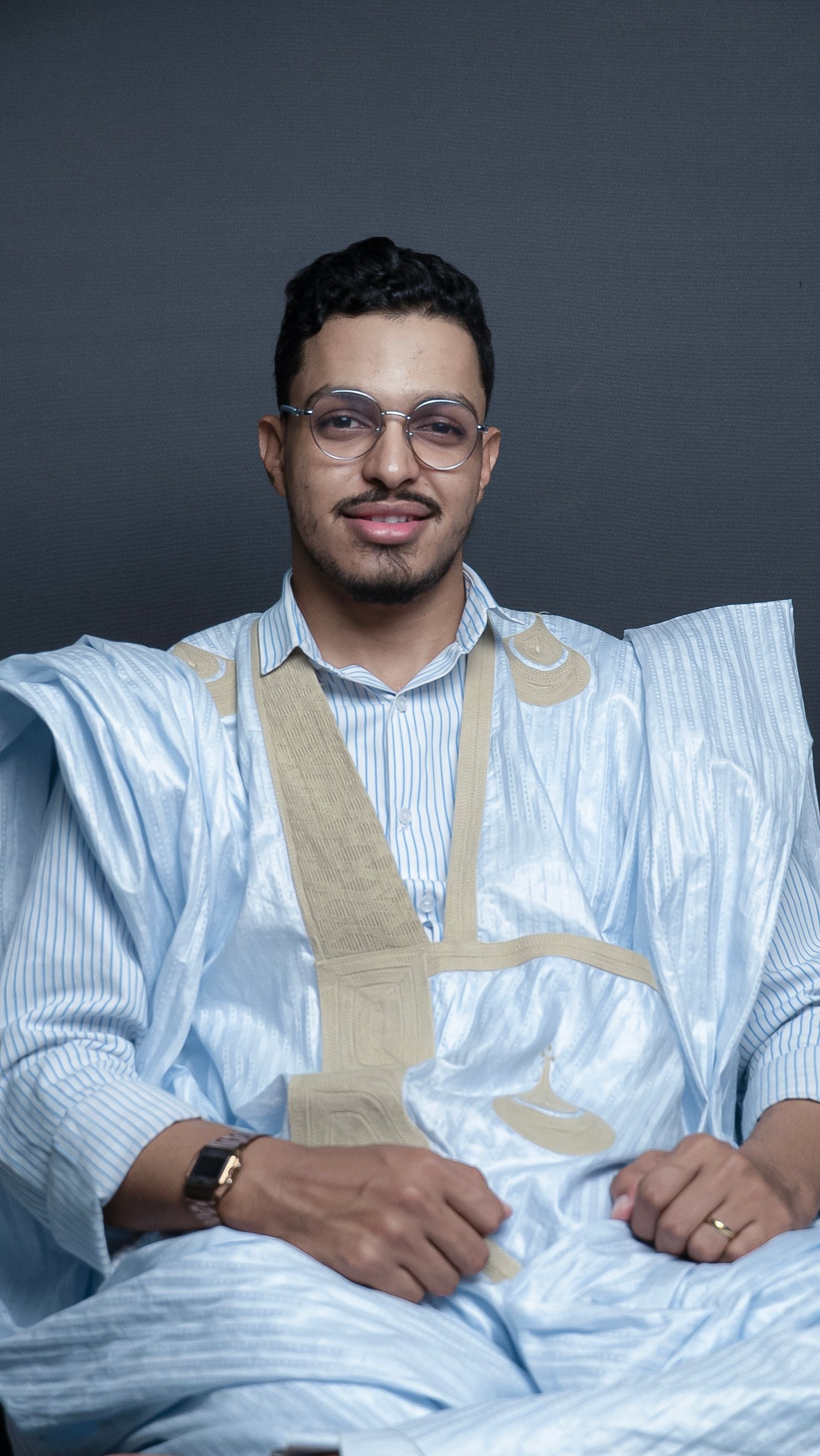 Portrait of a man in a traditional robe, wearing eyeglasses, sitting confidently.
