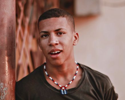 Close-up portrait of a young man leaning on a wooden structure wearing a colorful beaded necklace.