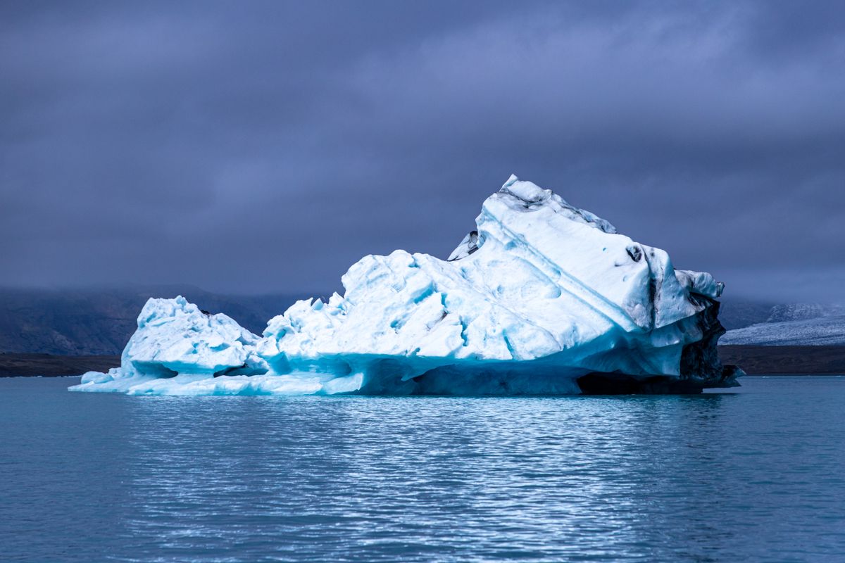 A large iceberg floating in cold Arctic-region waters under a moody sky with no people in the frame, a real photo chosen because drifting ice is what sets the story of Below in motion