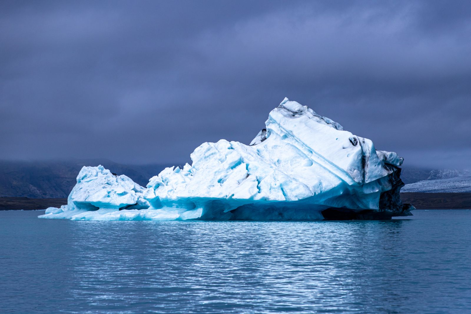 A large iceberg floating in cold Arctic-region waters under a moody sky with no people in the frame, a real photo chosen because drifting ice is what sets the story of Below in motion