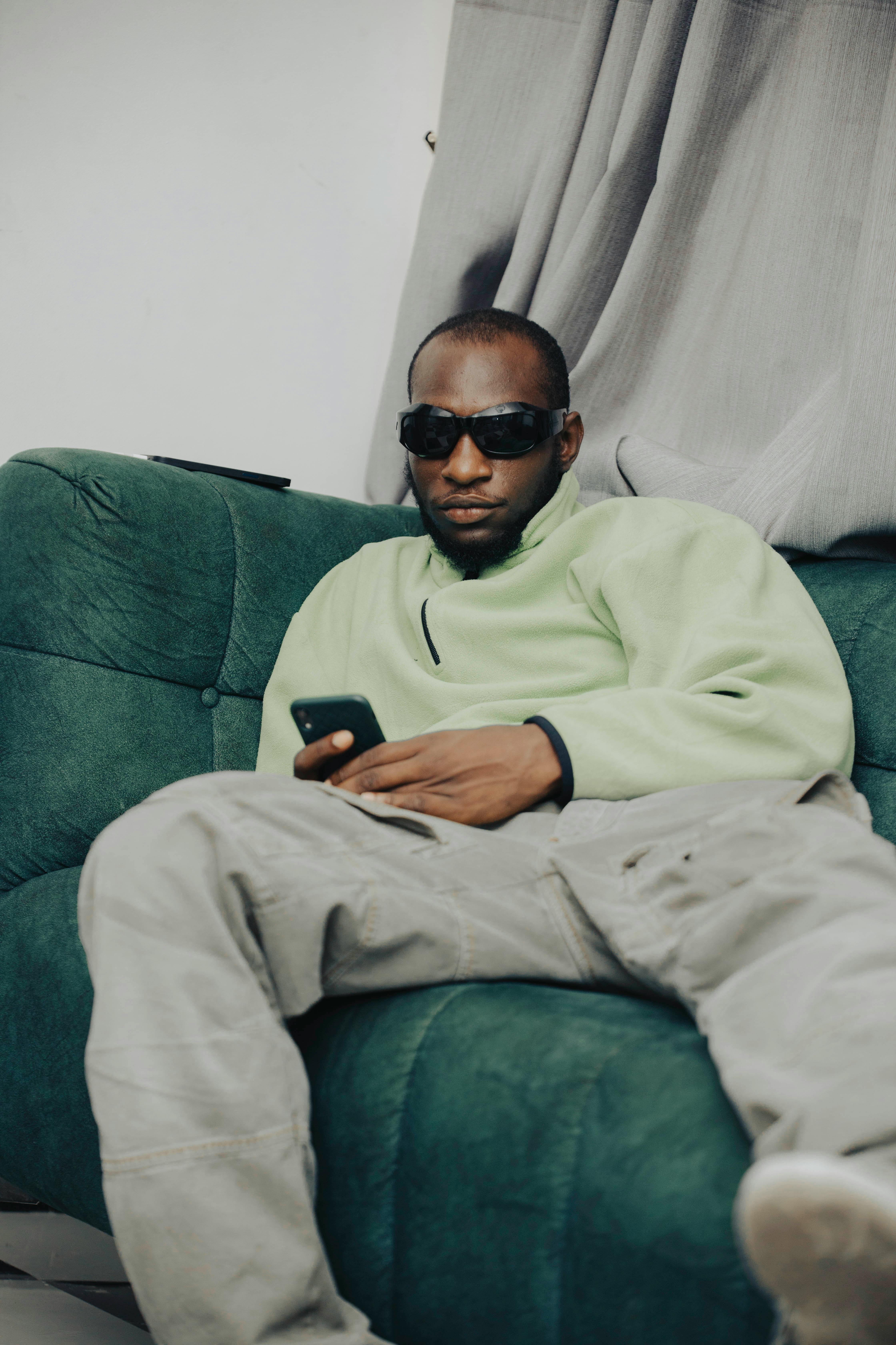 Free Man in sunglasses relaxes on green sofa using smartphone indoors. Stock Photo