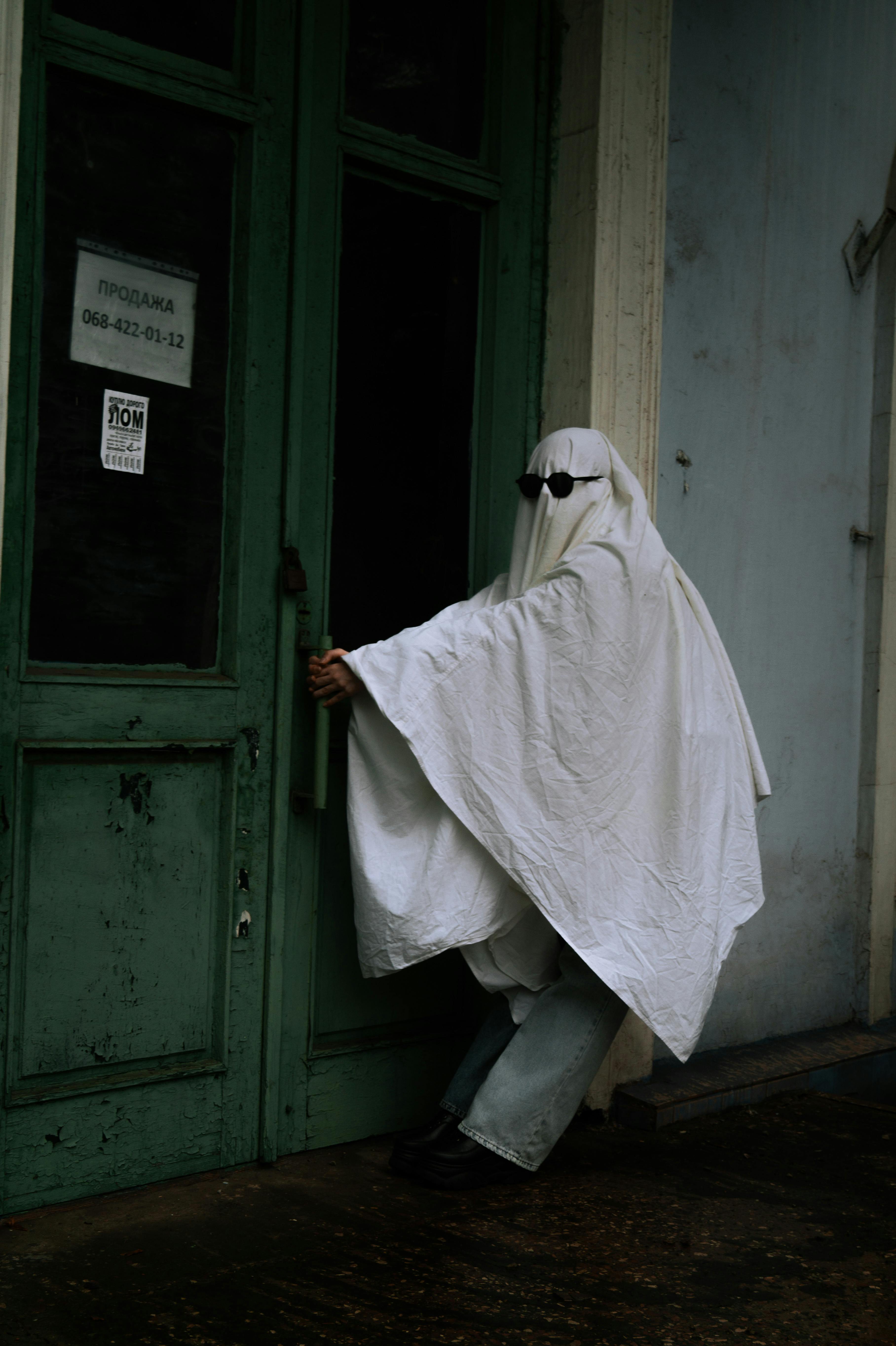 A person in ghost costume and sunglasses tries to open a green door in an urban setting.