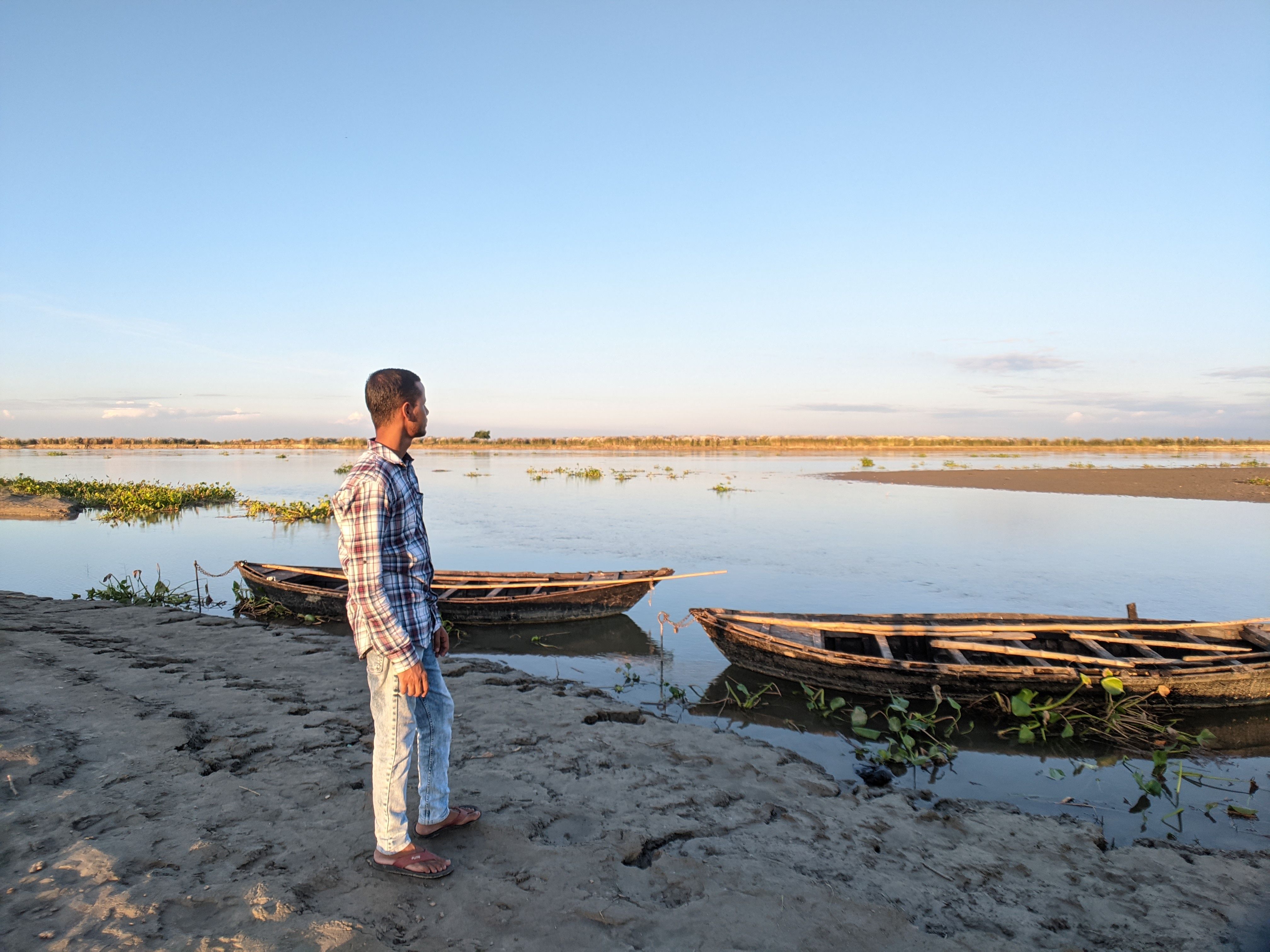 A man stands on a riverbank, observing wooden boats in a tranquil rural setting.